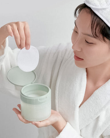 Person holding a green skincare container with a white lid, wearing a white robe and towel.