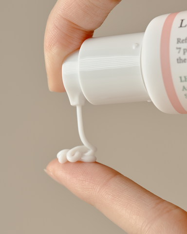 Hand holding a white bottle with pink label, squeezing cream onto a finger against a neutral background