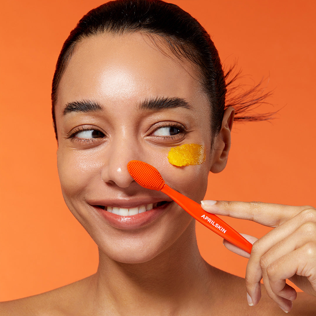Woman applying a yellow cream to her face with an orange applicator against an orange background