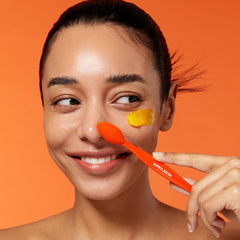 Woman applying a yellow cream to her face with an orange applicator against an orange background