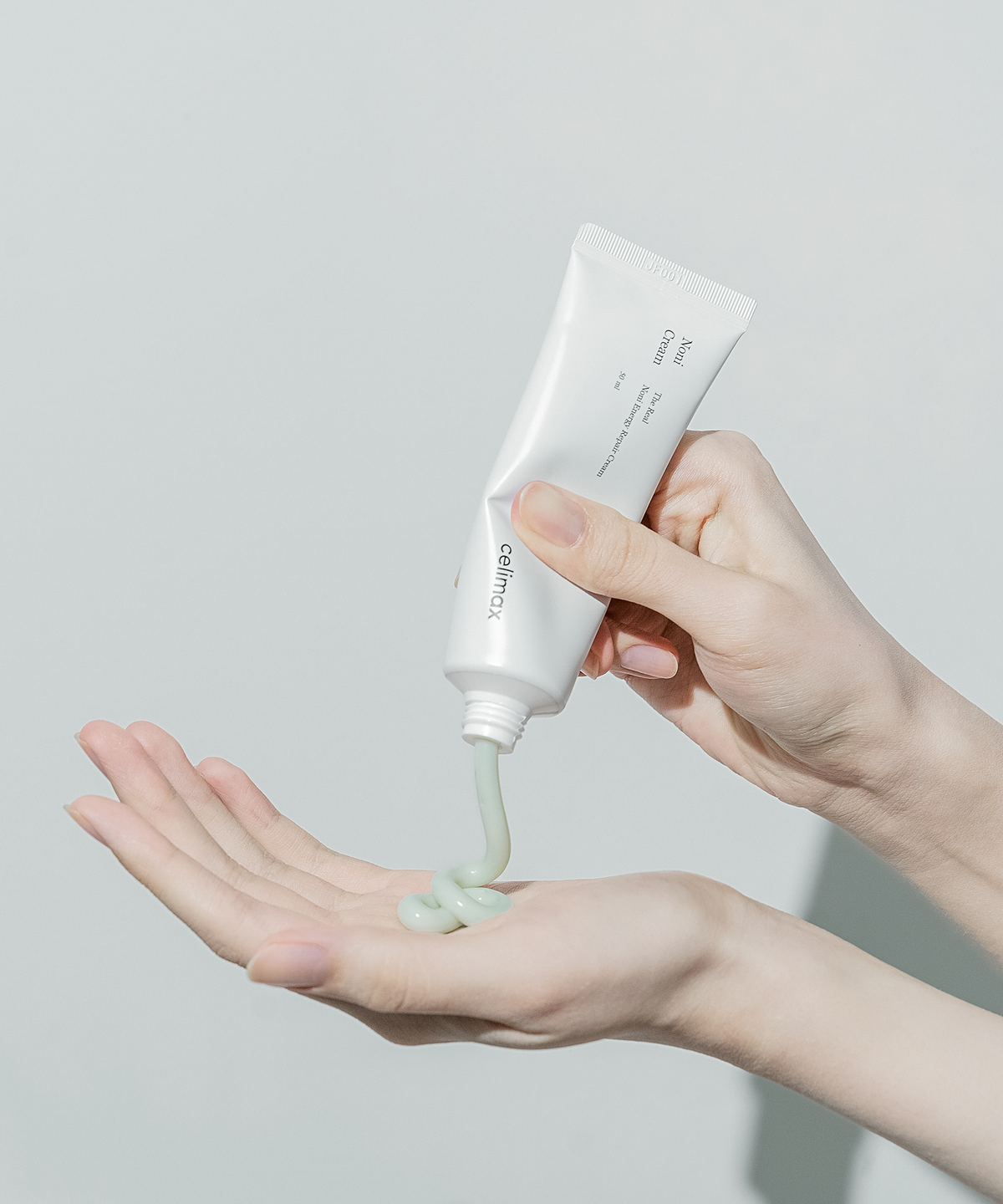 Hand holding a white tube of skincare product with green gel being squeezed onto another hand against a light gray background.