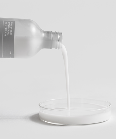 Bottle pouring white liquid into a small round container on a light gray background