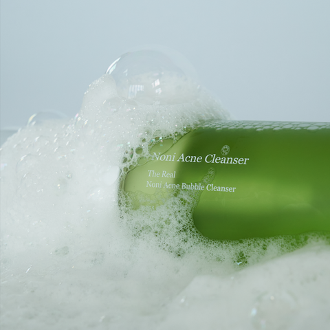 Green bottle of Nomi Acne Cleanser surrounded by bubbles on a light gray background