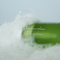 Green bottle of Nomi Acne Cleanser surrounded by bubbles on a light gray background