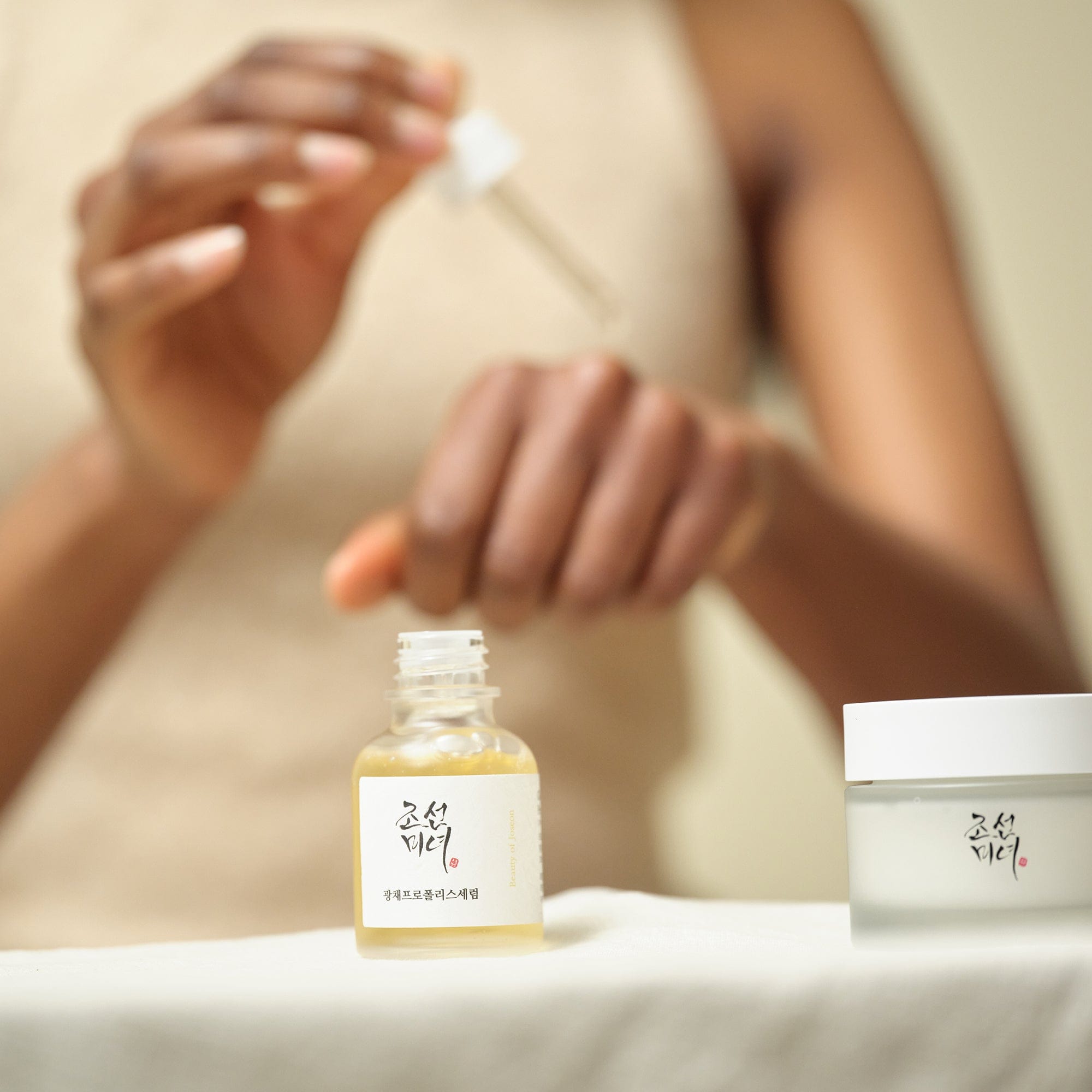Person applying a skincare product with a dropper, next to a bottle and jar of skincare items.