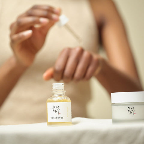 Person applying a skincare product with a dropper, next to a bottle and jar of skincare items.