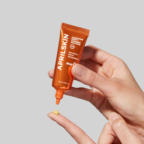 Hand holding an orange Aprilskin skincare product with a small amount on a finger against a light gray background