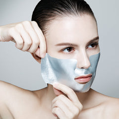 Woman applying a silver facial mask on a plain background
