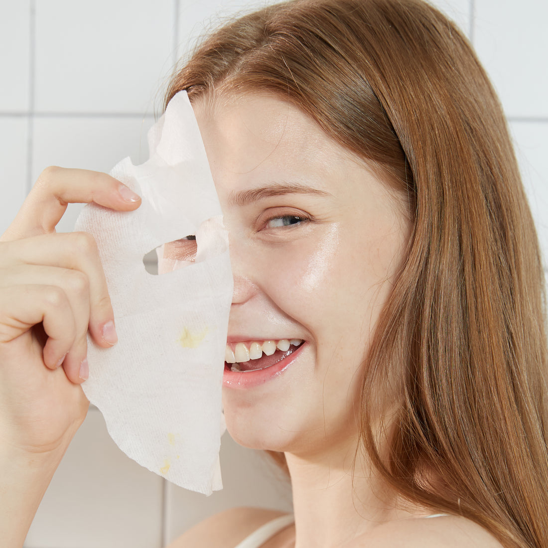 Woman holding a sheet mask up to her face with a white tiled background