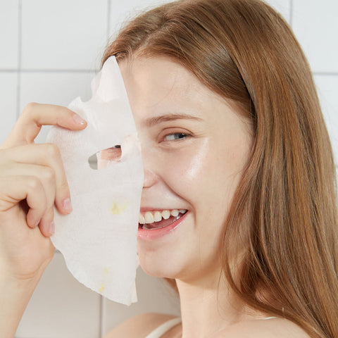 Woman holding a sheet mask up to her face with a white tiled background