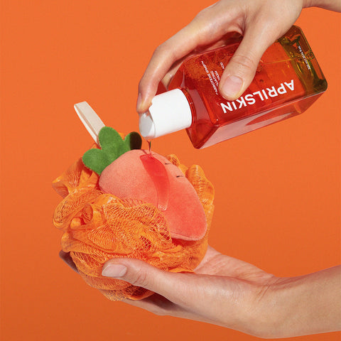 Hand holding a red bottle labeled 'APRILSKIN' over an orange bath pouf with a peach design on an orange background.