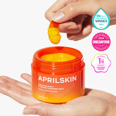 Orange jar of APRILSKIN cleansing balm with a hand holding a yellow scrub, on a white background.