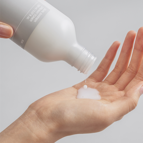 Person pouring a white cream from a bottle onto their palm against a light gray background