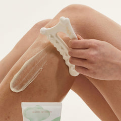 Person using a white exfoliating tool on their knee with a small container of cream below.